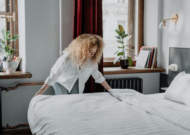 A woman with curly hair making a bed in a bright, cozy bedroom with plants and natural light.