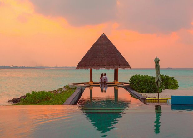 A couple enjoying a romantic moment by the sea and pool under a beach cabana during sunset.